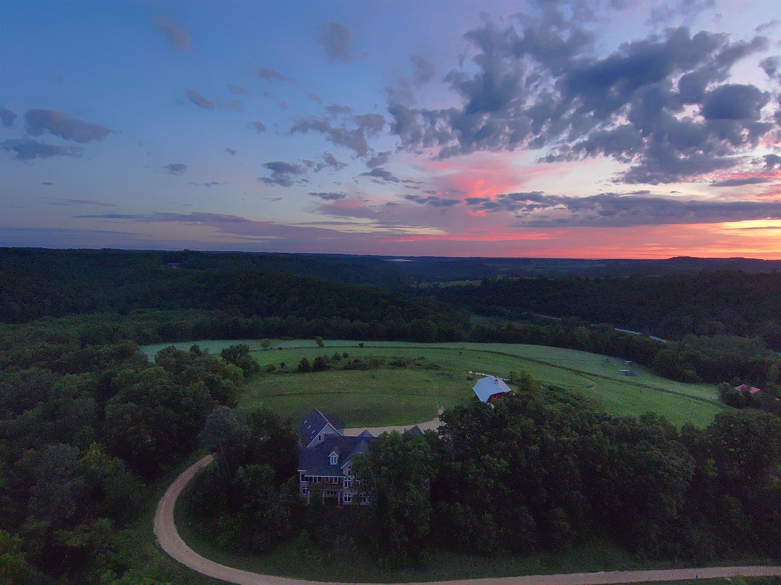 This aerial perspective captures a sprawling rural estate at sunset, featuring a multi-story home nestled among mature trees and a prominent red barn in the distance. A winding gravel driveway leads through the lush, rolling green landscape, emphasizing the property's expansive acreage and serene, secluded setting. The dramatic sky, painted with hues of pink and deep blue, provides a picturesque backdrop for this peaceful countryside retreat.