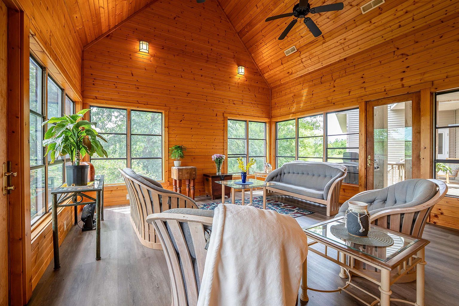 This bright and airy sunroom features warm wood-paneled walls and a vaulted ceiling, creating a cozy, rustic atmosphere. The space is furnished with a comfortable seating arrangement, including a sofa and matching armchairs, centered around a glass-topped coffee table. Large windows surround the room, providing abundant natural light and views of the outdoors, while a ceiling fan and wall-mounted lighting fixtures complete the space.