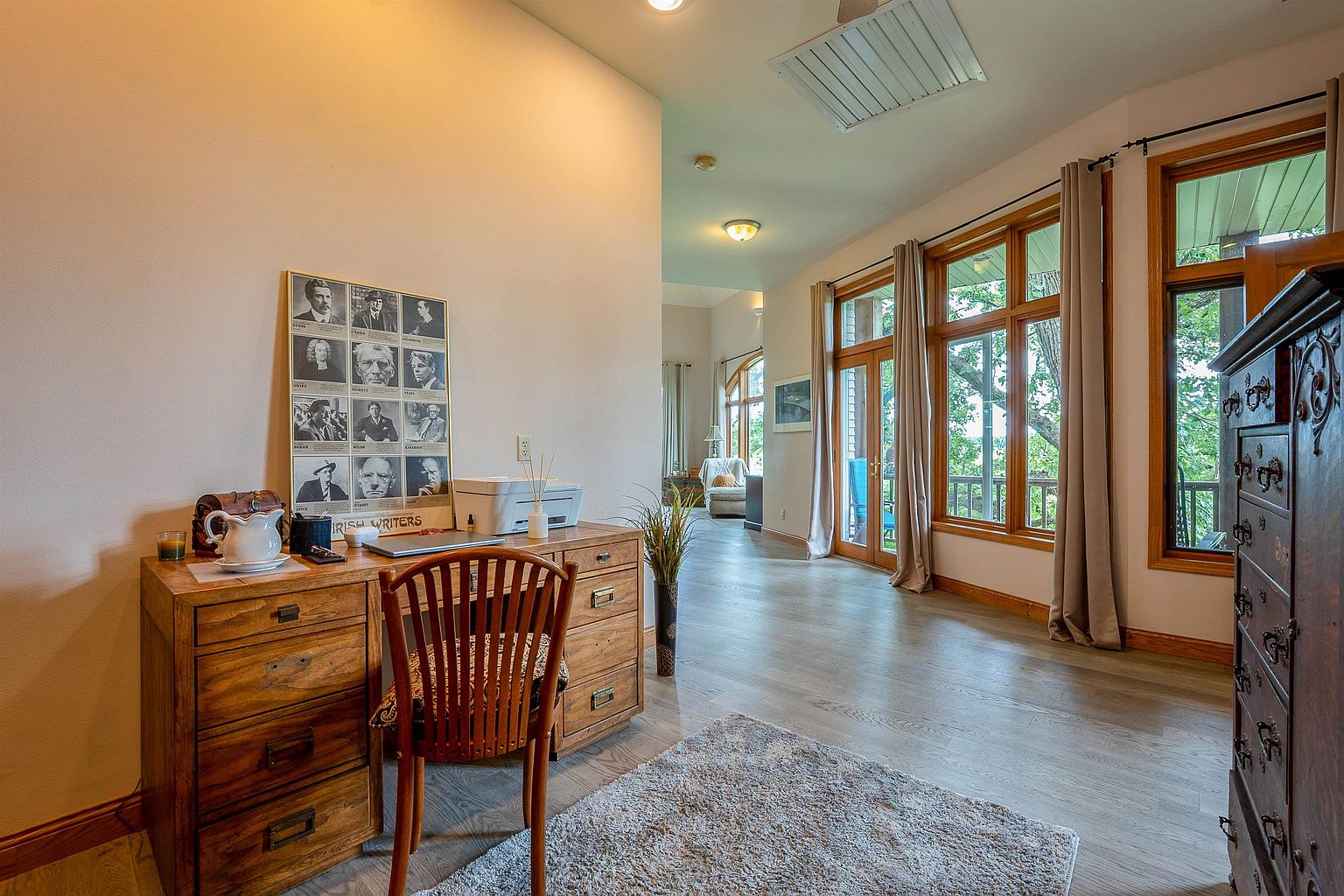This home office features a rustic wooden desk positioned against a wall adorned with a framed 'Irish Writers' poster, creating a scholarly and inviting atmosphere. The room is bathed in natural light from large windows that offer a view of the outdoors, complemented by light-toned wood flooring and a textured area rug. A hallway leads from the workspace into an adjacent living area, emphasizing an open and airy floor plan.