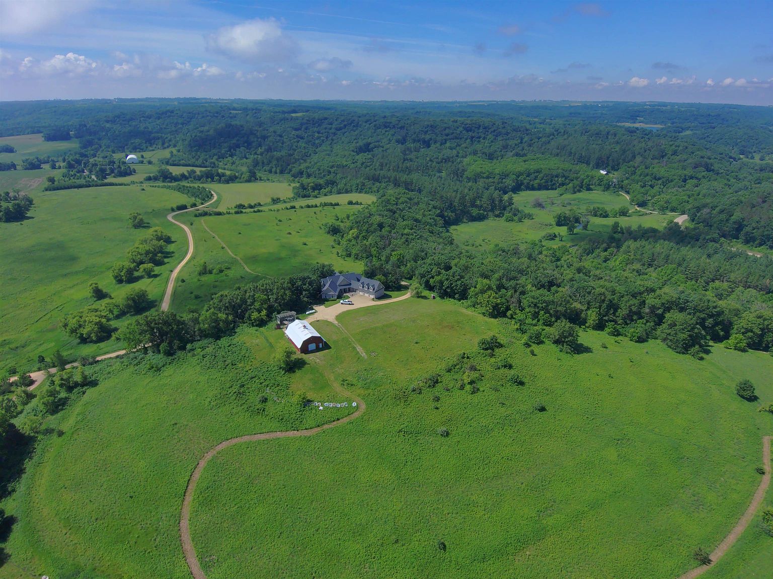 This high-angle aerial view captures a sprawling rural property set amidst a vast, rolling landscape of lush green fields and dense forests. The estate features a primary residence and a prominent red barn, connected by a gravel driveway that winds through the expansive grounds. The perspective emphasizes the property's seclusion, natural beauty, and significant acreage, offering a cinematic sense of scale and tranquility.
