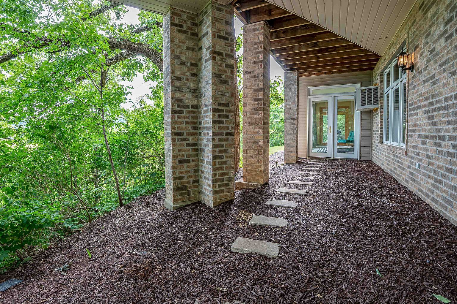 This covered patio area features a brick-clad structure with sturdy brick columns supporting the overhanging roof. A path of stone pavers leads across a bed of dark mulch toward a set of glass-paned double doors that provide access to the home's interior. The perspective captures the transition between the shaded outdoor living space and the lush, green foliage of the surrounding landscape.