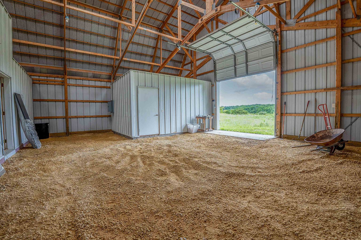 This image shows the interior of a large, utilitarian barn or workshop space with a gravel floor and exposed wooden truss ceiling. A white metal-clad storage room stands in the center, while a large overhead garage door is open, revealing a view of a grassy field and distant hills. The space is sparsely furnished with a wheelbarrow, some tools, and a trash bin, suggesting a functional, multi-purpose storage or workspace environment.
