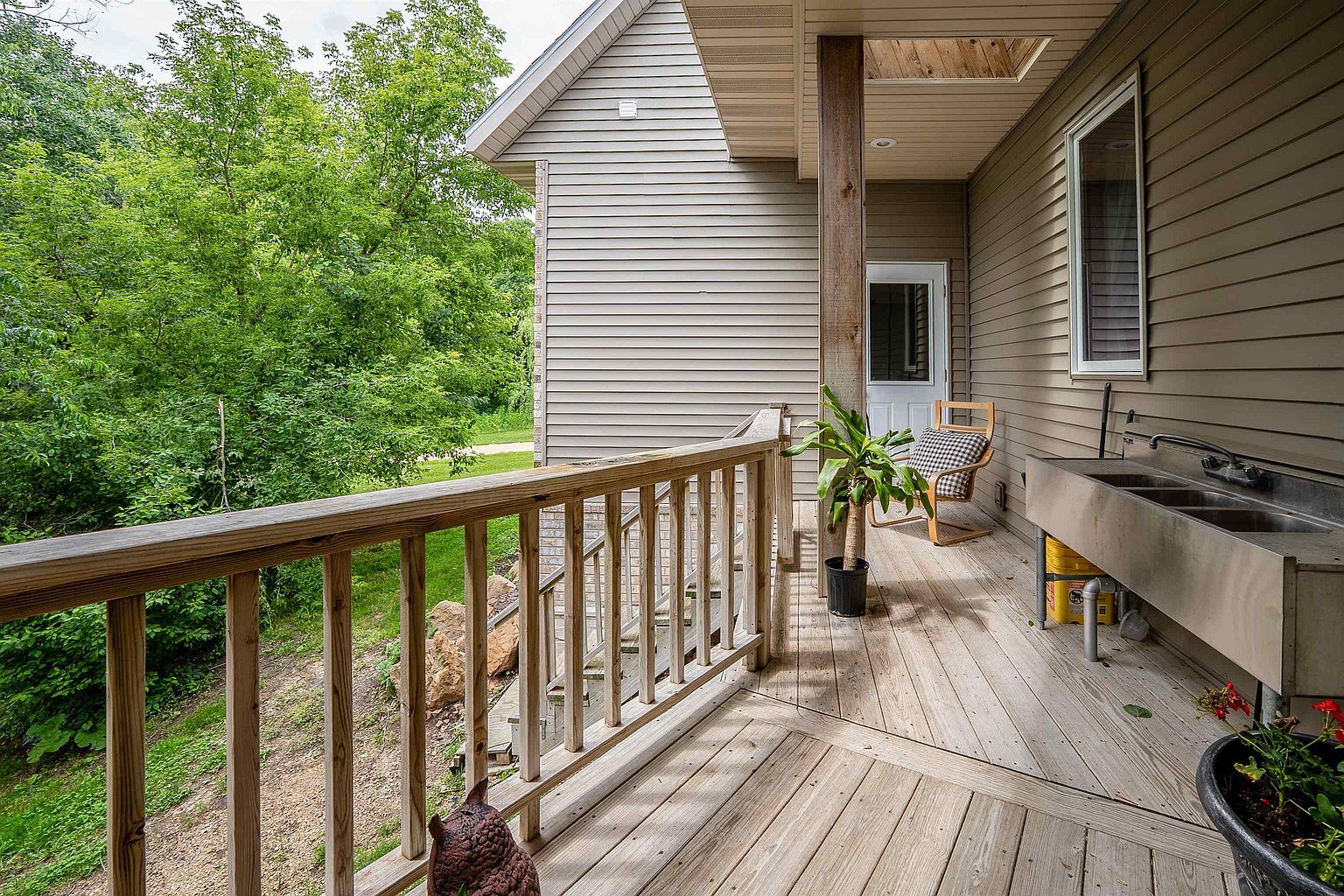 This elevated wooden deck offers a functional outdoor space featuring a stainless steel utility sink and a wooden chair with a checkered cushion. The deck is bordered by a simple wooden railing and overlooks a lush, green wooded area, providing a private and natural setting. The perspective captures the transition from the home's siding to the outdoor living area, emphasizing the connection to the surrounding landscape.