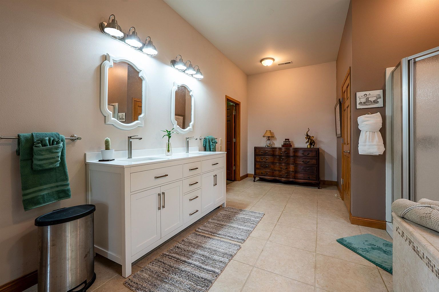 This spacious primary bathroom features a long white double vanity with dual mirrors and modern light fixtures. The room is finished with neutral-toned tile flooring, a dark wood dresser for additional storage, and a walk-in shower area. The overall aesthetic is clean and functional, offering a bright and well-organized space.
