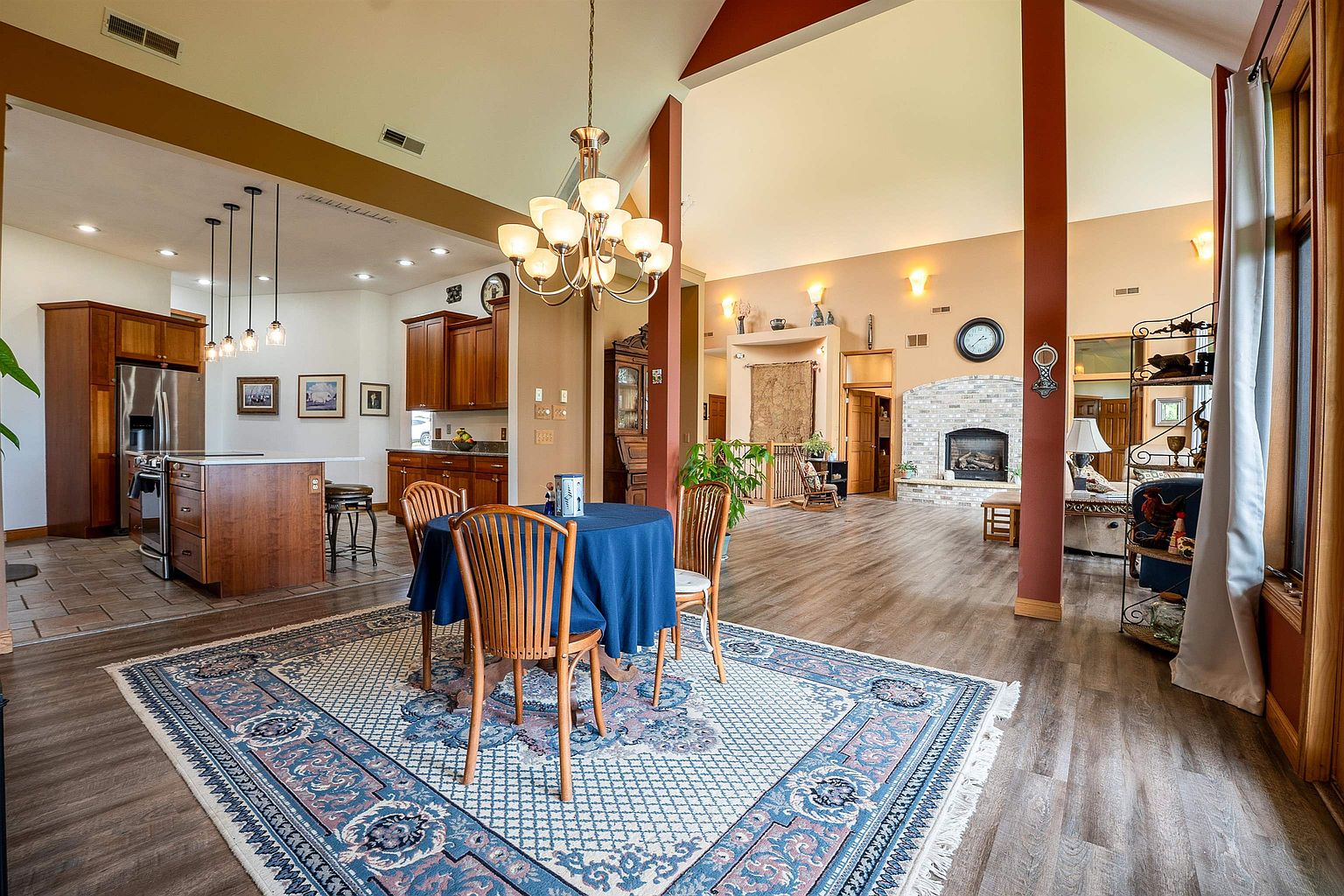 This open-concept living space features a central dining area with a round table and blue tablecloth, anchored by a large, ornate patterned rug. To the left, a spacious kitchen with wood cabinetry and a central island is visible, while the right side opens into a living area centered around a brick-surround fireplace. The room boasts high vaulted ceilings, warm wall tones, and a mix of wood-look flooring and tile, creating a cohesive and inviting atmosphere.