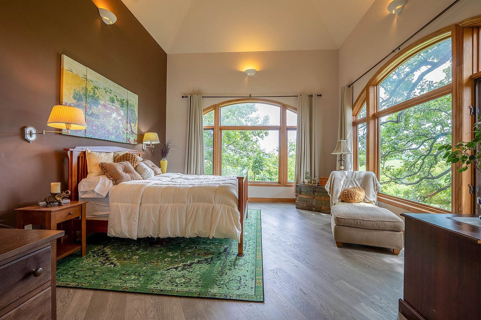 This spacious primary bedroom features a vaulted ceiling and large, arched windows that provide abundant natural light and scenic views of the surrounding trees. The room is anchored by a wooden bed frame topped with a white comforter and a vibrant green patterned area rug, complemented by a cozy chaise lounge positioned near the window. The warm brown accent wall behind the bed creates a sophisticated focal point, while the light wood-toned flooring adds a modern, airy feel to the space.