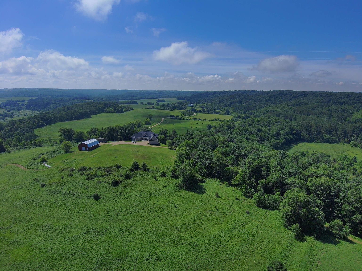 This wide-angle aerial perspective captures a sprawling rural estate nestled within a lush, rolling landscape of green fields and dense forest. The property features a primary residence and a distinct red barn, both situated on a gentle rise that overlooks the expansive, undulating terrain. The bright blue sky with scattered clouds enhances the serene and secluded atmosphere of this expansive countryside setting.