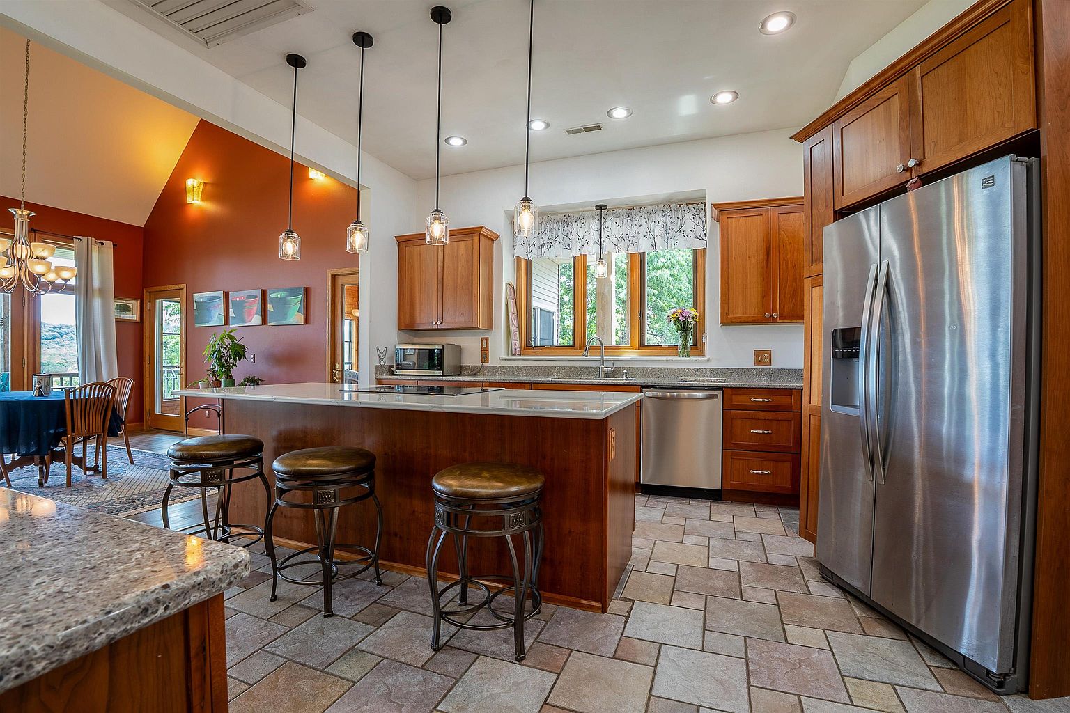 This spacious kitchen features warm-toned wood cabinetry, a large central island with stone countertops, and three pendant lights hanging above. The room is equipped with stainless steel appliances, including a refrigerator and dishwasher, and is finished with patterned tile flooring. A wide window over the sink provides natural light, while the adjacent dining area is visible through an open transition.
