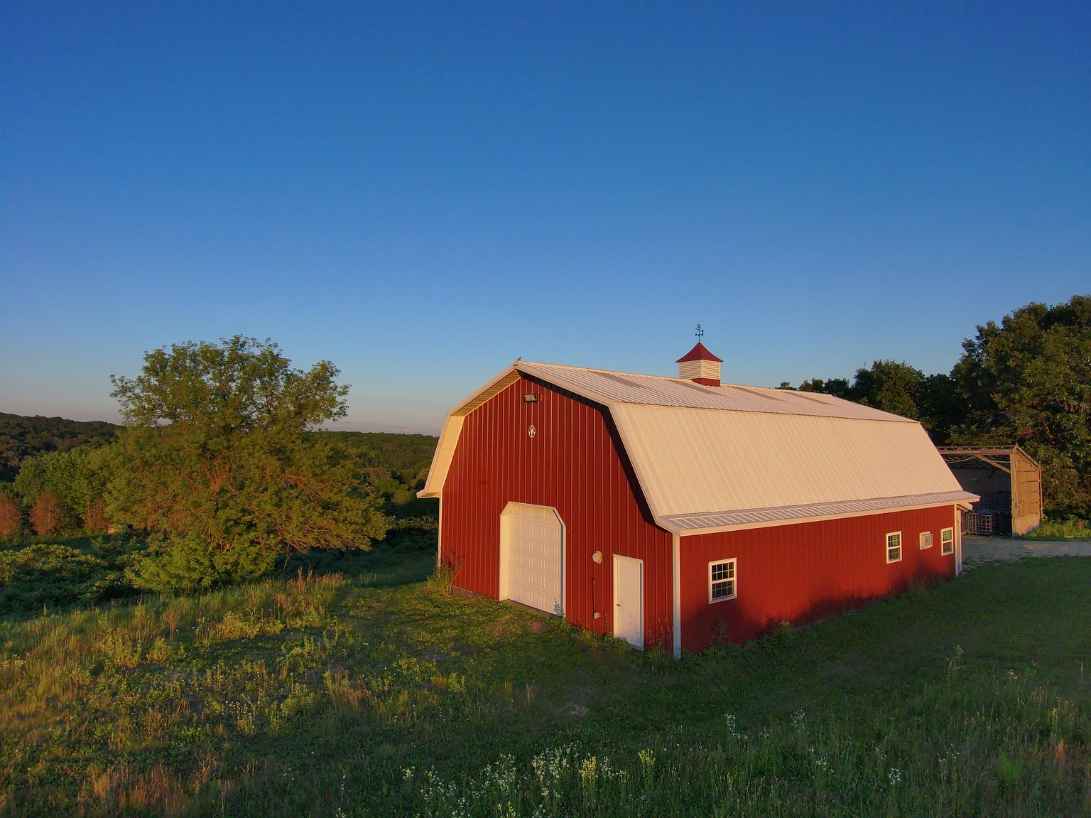 This aerial perspective captures a classic red barn with a white metal roof, set against a backdrop of rolling hills and lush greenery during the golden hour. The structure features a prominent white garage door, a side entry door, and several windows, all surrounded by a sprawling grassy landscape. The warm, low-angle sunlight highlights the barn's textures and creates a serene, pastoral atmosphere.