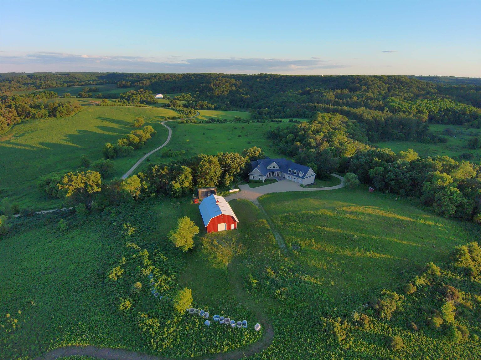 This high-angle aerial perspective captures a sprawling rural estate set amidst rolling green hills and dense forest. The property features a large, multi-gabled residence with an attached garage, complemented by a prominent red barn situated on the grassy slope in the foreground. A winding gravel driveway connects the structures, emphasizing the secluded and expansive nature of the landscape.
