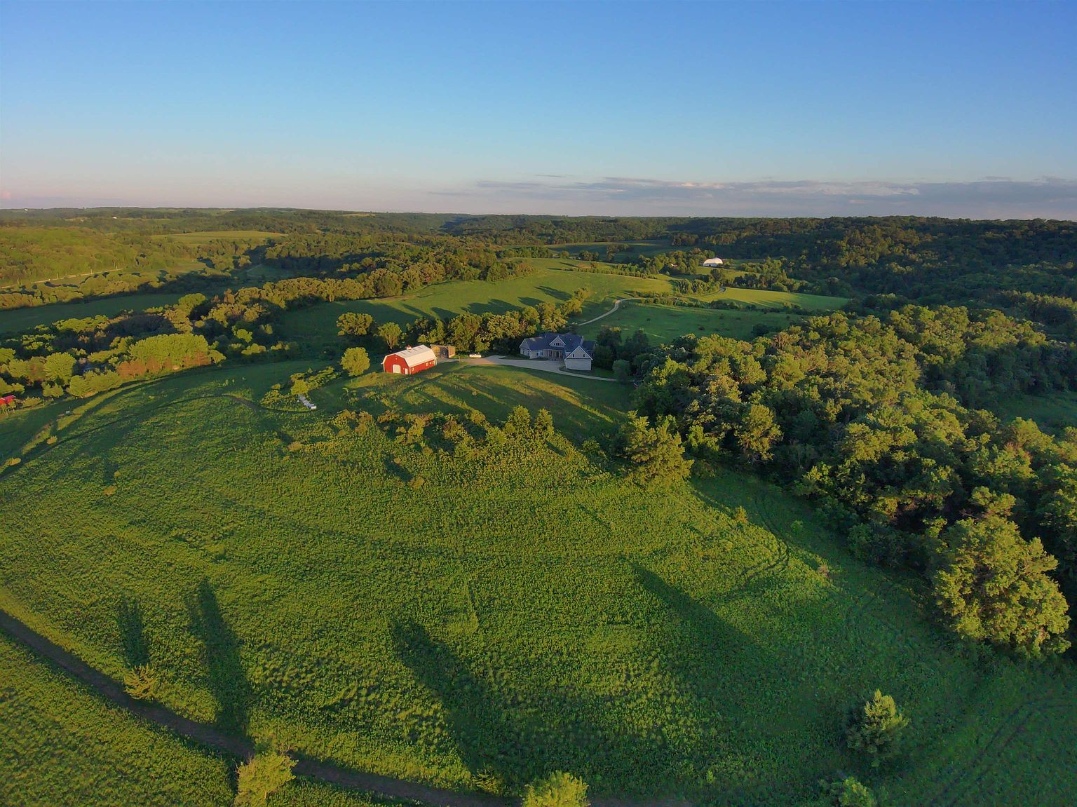 This high-angle aerial view captures a sprawling rural property set amidst rolling green hills and dense patches of forest. The estate features a prominent red barn and a residential home situated on a grassy ridge, with a winding driveway leading through the landscape. The golden hour lighting casts long shadows, emphasizing the vast, serene topography of the surrounding countryside.