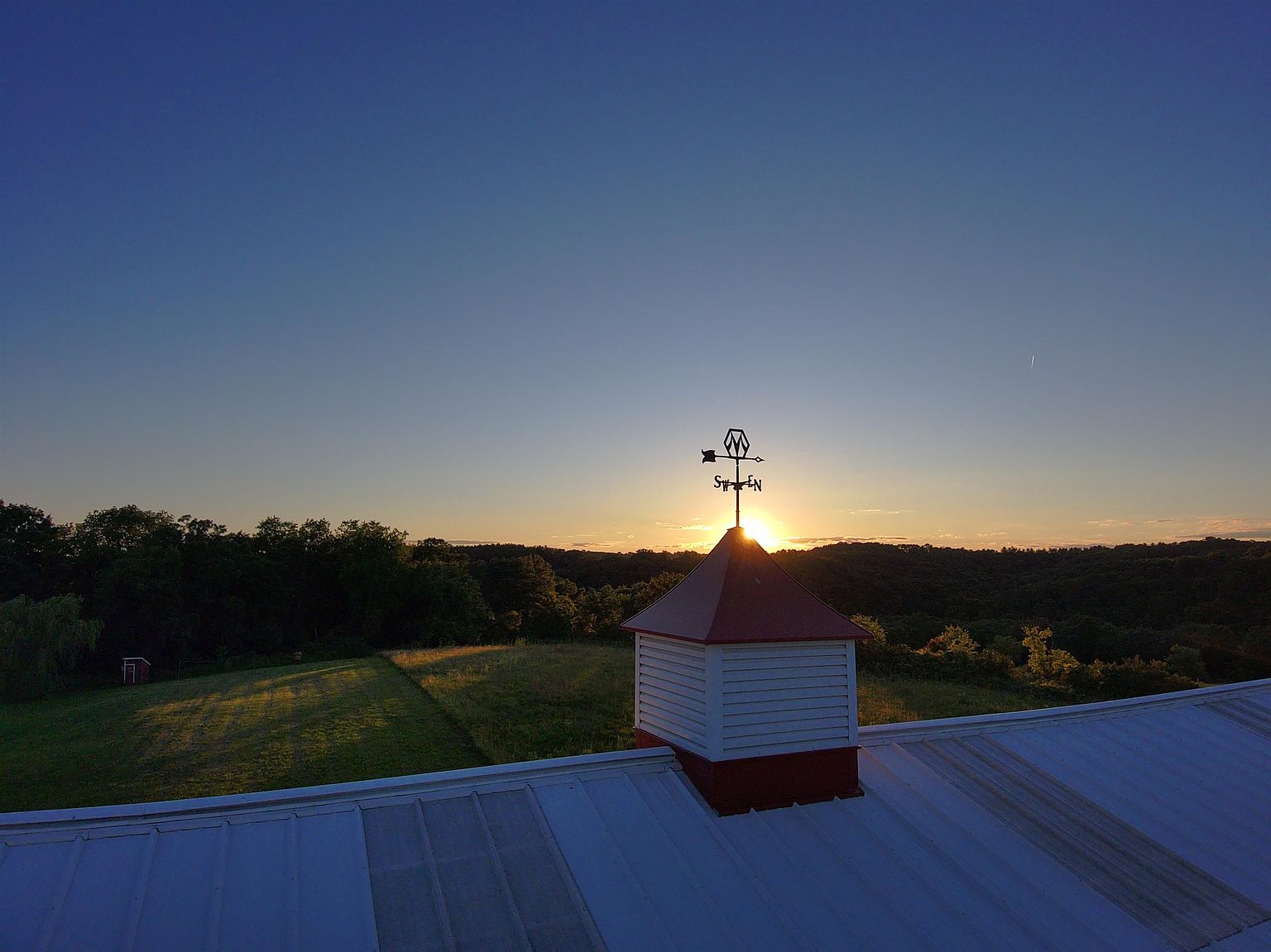 This elevated perspective captures a serene sunset over a rural landscape, focusing on the cupola and weather vane atop a metal-roofed structure. The composition highlights the silhouette of the weather vane against the glowing sun, framed by a vast, clear blue sky and rolling hills in the distance. The scene evokes a peaceful, pastoral atmosphere, emphasizing the property's connection to its natural surroundings.