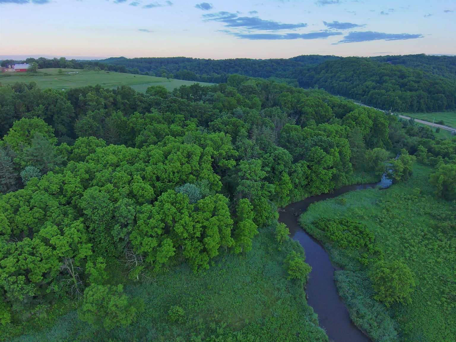 This aerial perspective captures a sprawling rural landscape featuring a winding creek that meanders through lush green meadows and dense forest. In the distance, a small red barn sits atop a rolling hill, emphasizing the expansive and tranquil nature of the property. The scene is bathed in the soft, golden light of late afternoon, highlighting the natural beauty and privacy of the terrain.