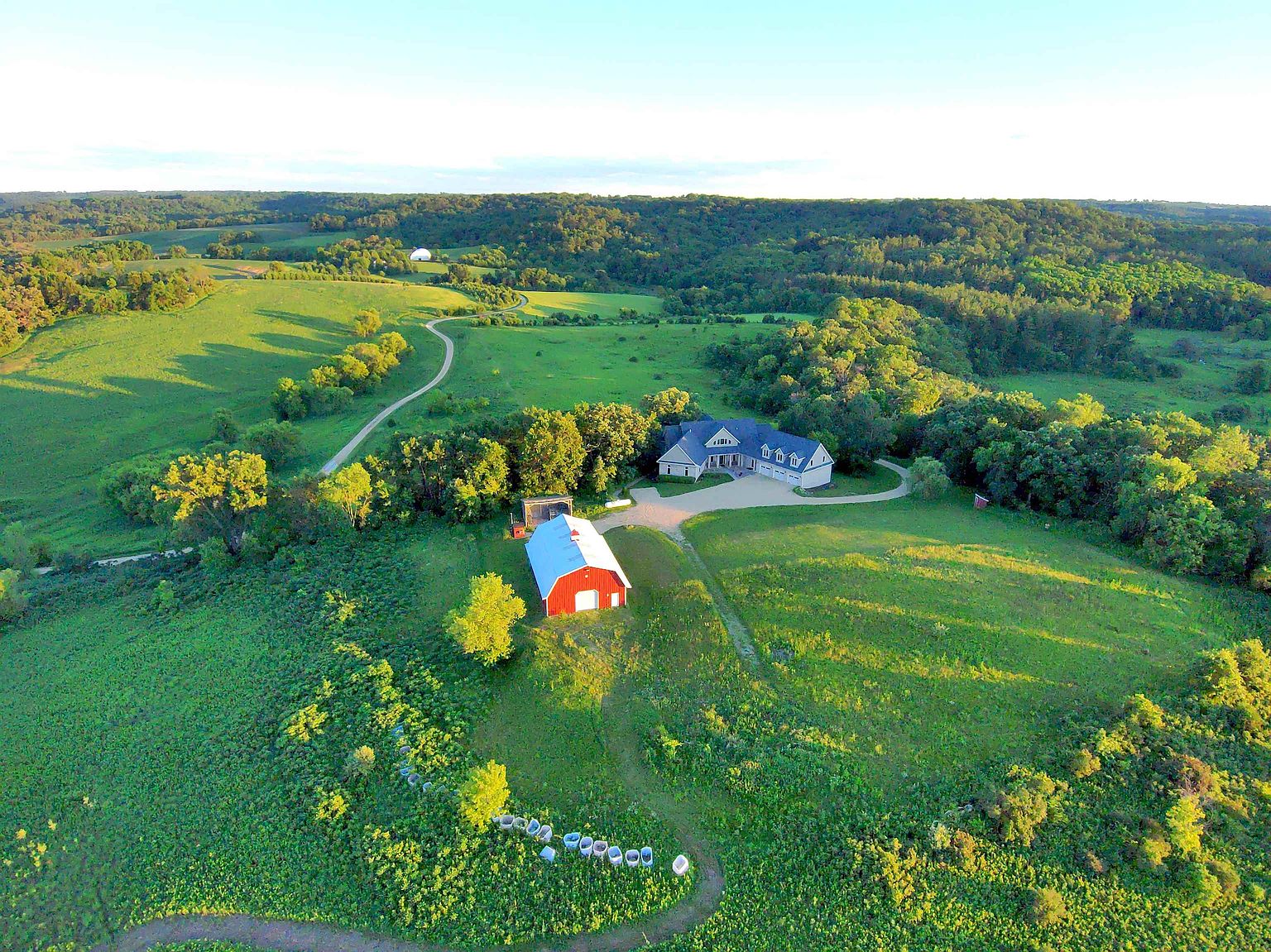 This high-angle aerial view captures a sprawling rural estate set amidst rolling green hills and dense forest. The property features a large, multi-gabled residence with a paved driveway, accompanied by a prominent red barn with a metal roof situated on a grassy knoll. A winding dirt path meanders through the landscape, emphasizing the expansive and secluded nature of this countryside setting.