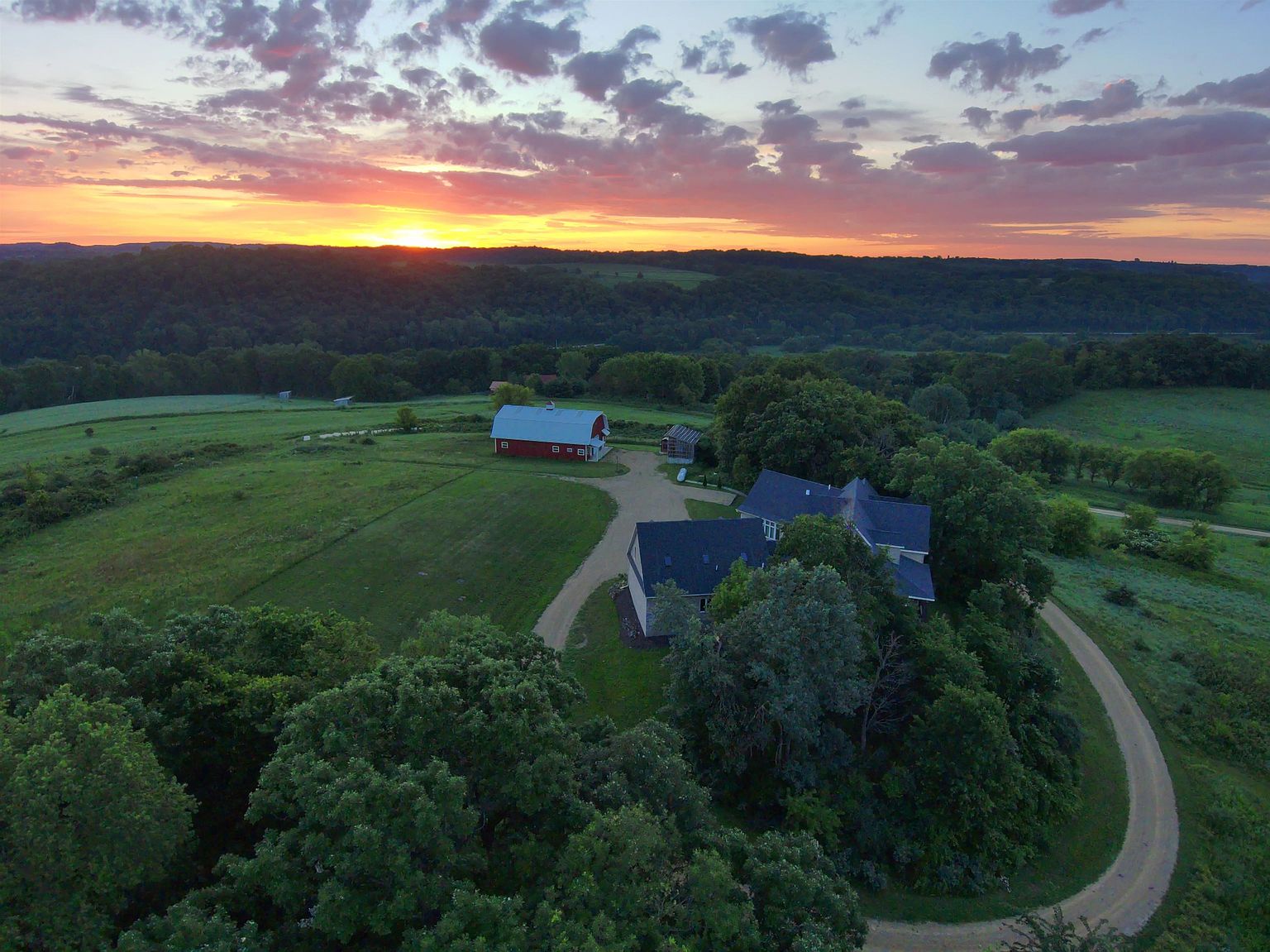 This stunning aerial perspective captures a sprawling rural estate at sunset, featuring a large multi-gabled residence nestled among mature trees. A gravel driveway winds through the lush green landscape, connecting the main house to a classic red barn in the distance. The dramatic sky, filled with vibrant orange and purple hues, provides a picturesque backdrop for this serene and expansive property.