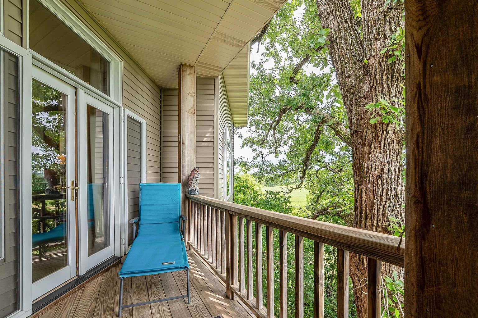 This elevated wooden deck offers a serene outdoor retreat, featuring a bright blue lounge chair positioned for relaxation. The space is framed by a wooden railing and a large, mature tree that provides natural shade and a scenic view of the surrounding greenery. Large glass doors lead from the deck into the home, creating a seamless transition between indoor and outdoor living areas.