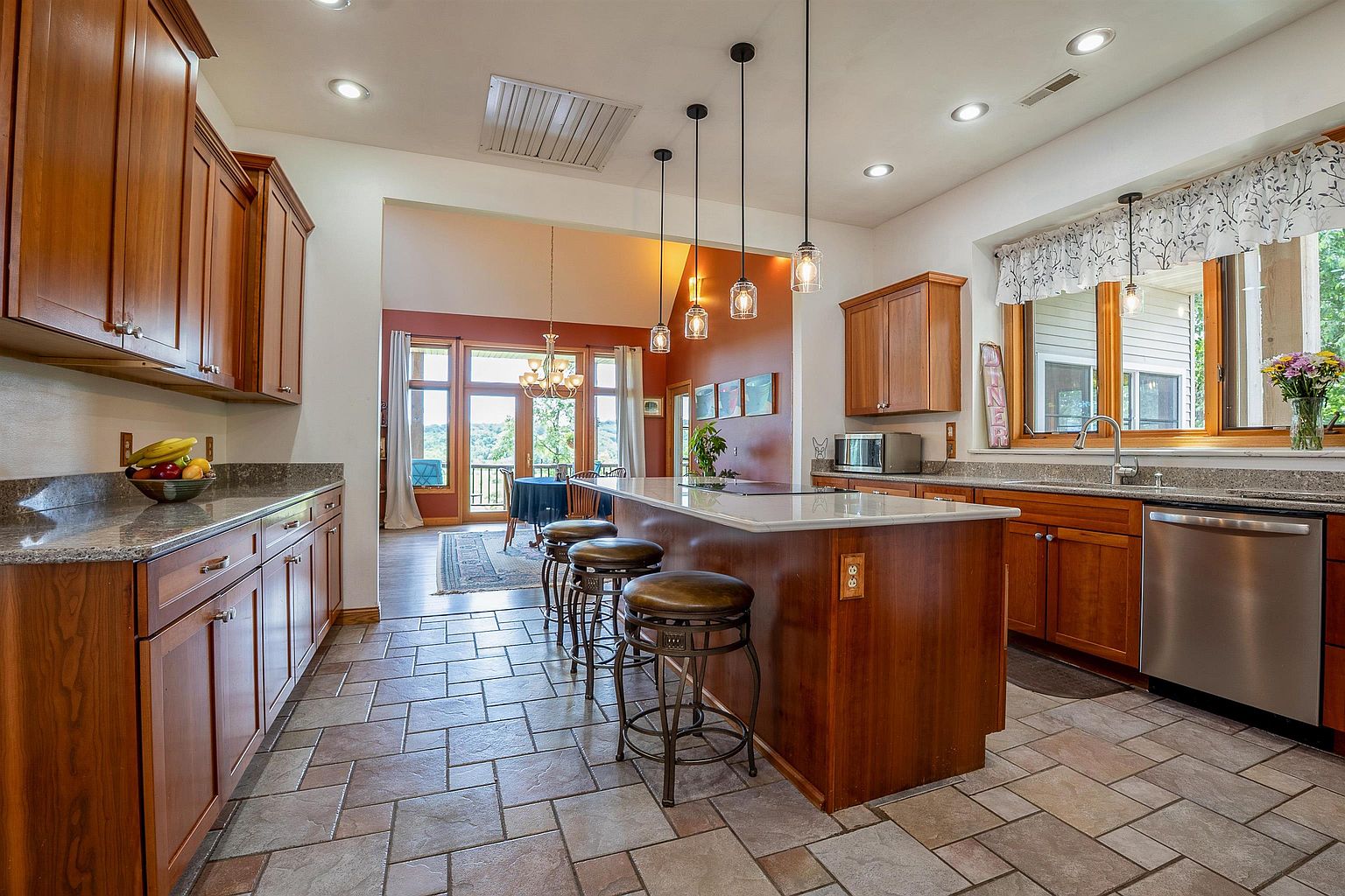This spacious kitchen features warm-toned wood cabinetry, a large central island with stone countertops, and three pendant lights hanging above. The room is finished with patterned tile flooring and opens into a dining area with large windows that provide a view of the outdoors. The overall aesthetic is traditional and inviting, with a functional layout that connects the cooking space to the rest of the home.