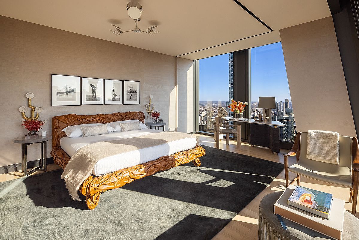 This is a luxurious primary bedroom featuring a unique, sculpted wooden bed frame with a plush white duvet and throw. The room boasts floor-to-ceiling windows offering stunning city views, complemented by a modern desk area and a stylish armchair. The overall aesthetic is sophisticated and contemporary, emphasizing comfort and elegance.