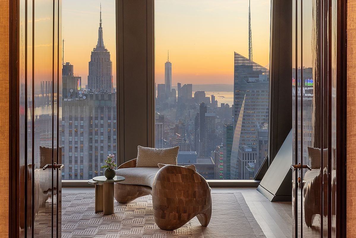 This interior shot showcases a luxurious living room with breathtaking city views, including the Empire State Building and One World Trade Center. The room features stylish furniture, including a unique wooden chair and a modern sofa, complemented by a small side table with flowers. The large windows and elegant flooring enhance the sophisticated ambiance, creating an inviting and upscale living space.