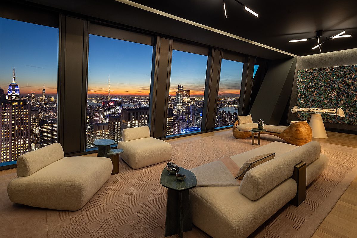 This is a luxurious living room with floor-to-ceiling windows offering a stunning cityscape view at dusk. The room features modern, plush furniture, including a curved sofa and lounge chairs, arranged on a patterned rug. The dark ceiling and contemporary lighting fixtures add to the sophisticated ambiance, creating an inviting and stylish space.