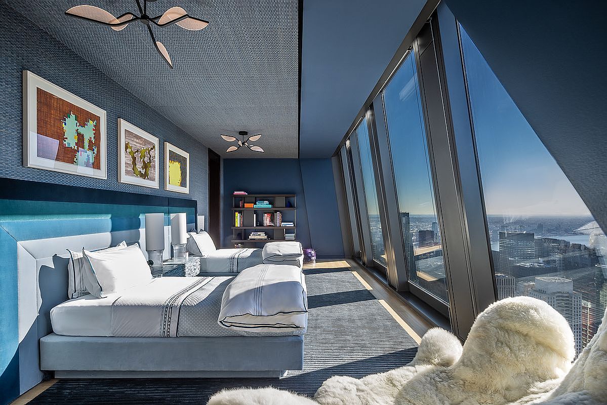 This is a modern bedroom featuring two twin beds with light blue upholstered headboards and crisp white linens. The room is decorated in shades of blue, with textured wallpaper and artwork adding visual interest. Large windows offer stunning city views, and a plush white rug adds a touch of luxury. The perspective is from the foot of one of the beds, showcasing the room's layout and the expansive cityscape beyond.