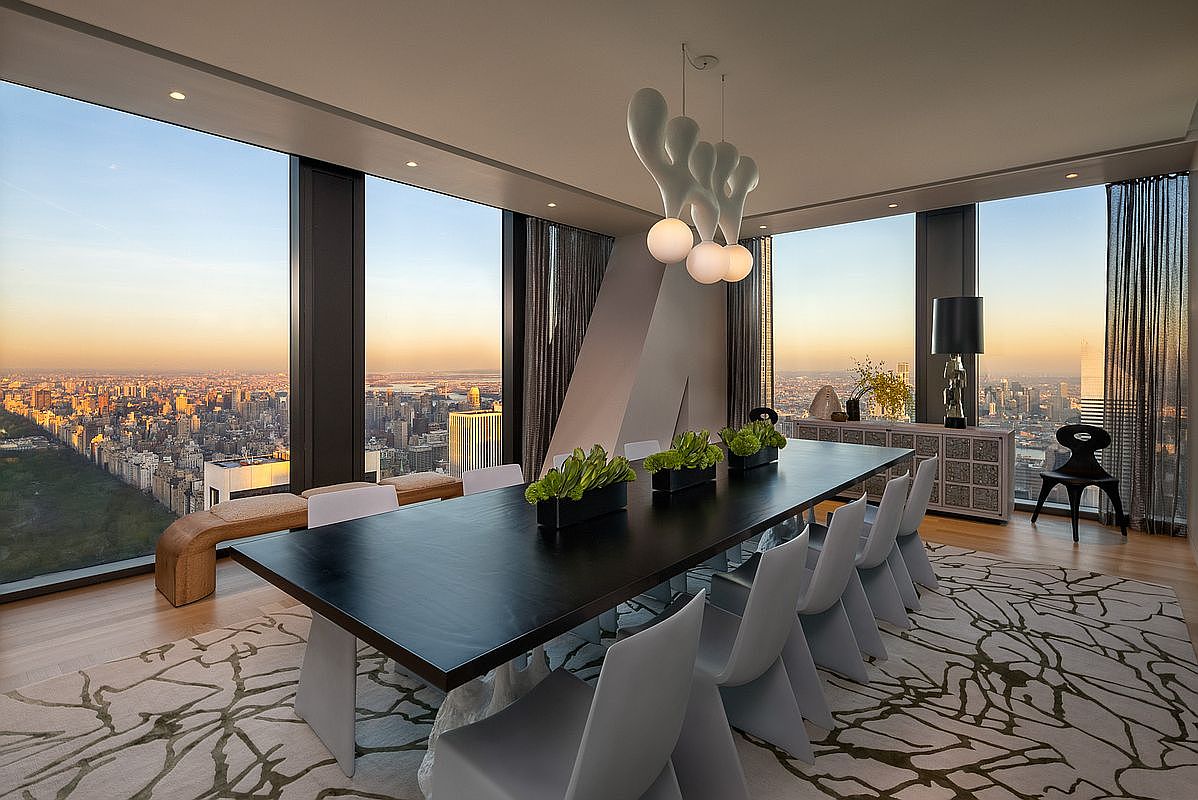 This is a luxurious dining room with a large, dark wood table surrounded by modern, light gray chairs. The room features floor-to-ceiling windows offering a panoramic city view at sunset, complemented by a unique, sculptural chandelier. A patterned rug adds texture to the space, creating an elegant and sophisticated atmosphere.