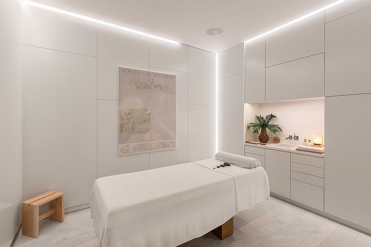 The image showcases a serene and minimalist spa treatment room. A massage table with a white linen cover is the focal point, complemented by a wooden stool and an abstract painting on the wall. Built-in white cabinets and soft lighting create a calming and luxurious atmosphere, ideal for relaxation and wellness.