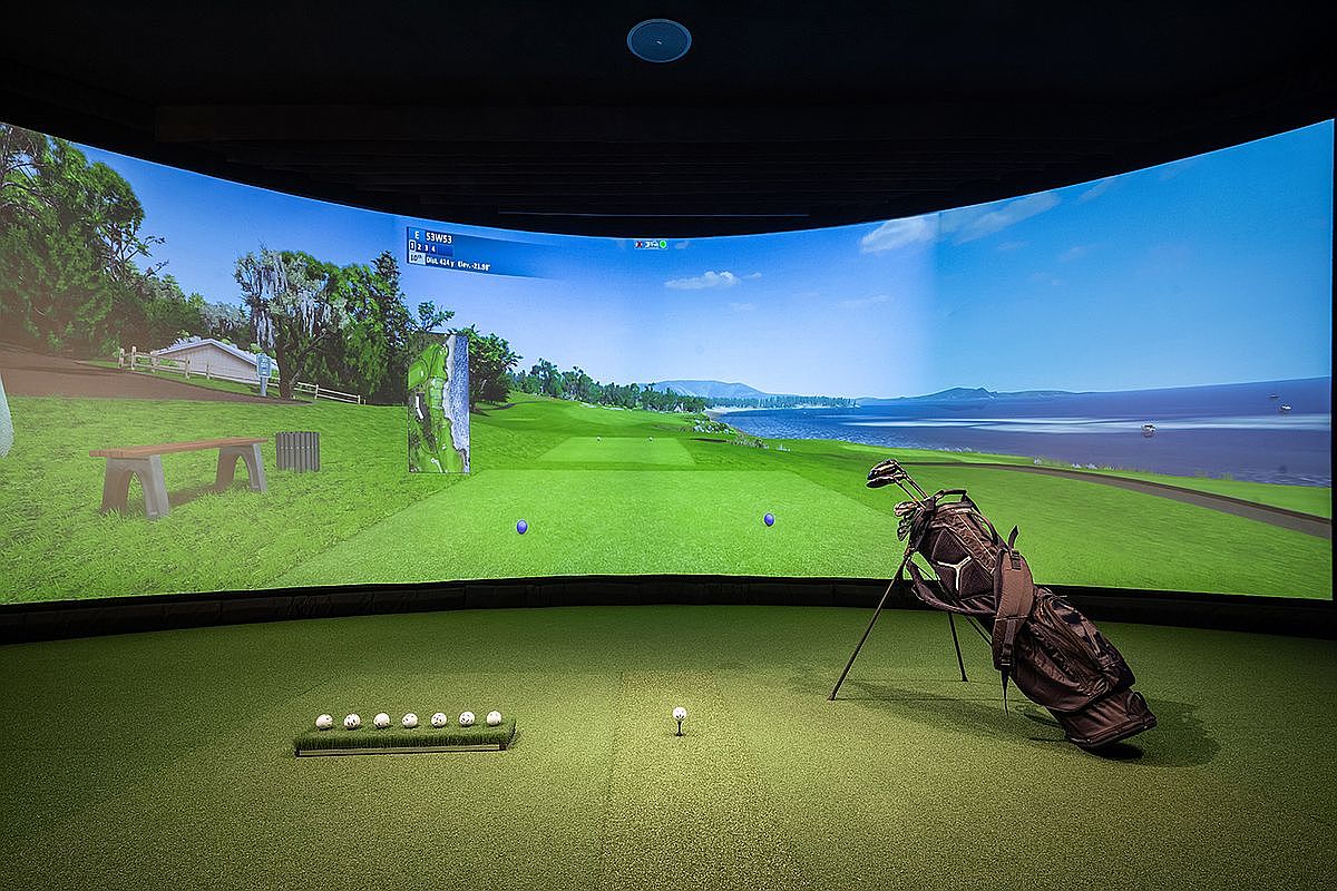 This image showcases an indoor golf simulator setup, featuring a large curved screen displaying a realistic golf course scene. The foreground includes a golf bag with clubs, a row of golf balls, and a tee, all set on artificial turf. The room is designed to replicate the experience of playing golf indoors, offering a unique amenity for a luxury home.