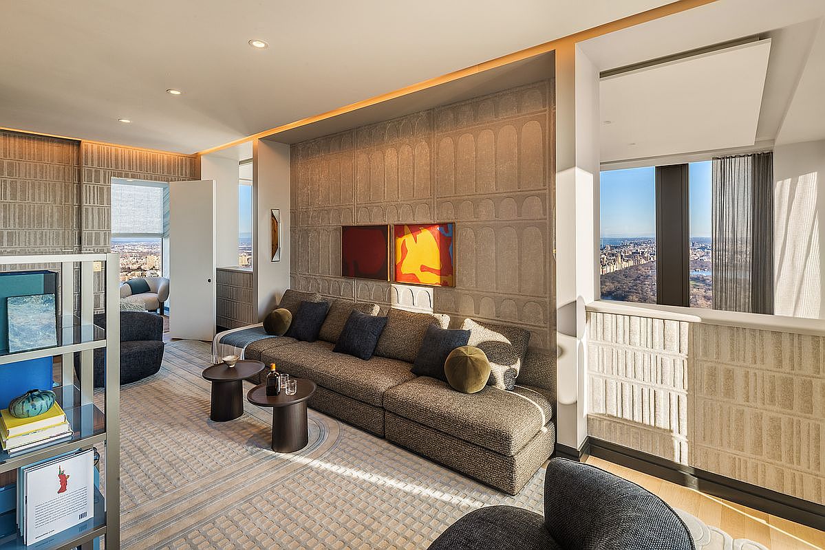 This is a stylish living room featuring a large, modular sofa with decorative pillows, complemented by two round side tables. The room is decorated with modern art and textured wall panels, creating a sophisticated and inviting atmosphere. Large windows offer a stunning city view, enhancing the luxurious feel of the space.