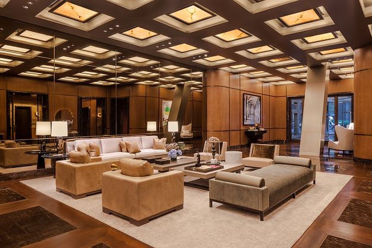 This is a luxurious living room featuring a neutral color palette with brown wood paneling and a coffered ceiling with recessed lighting. The room is furnished with plush sofas, armchairs, and a coffee table, all arranged on a large area rug. Mirrored walls enhance the sense of space and reflect the light, creating a bright and inviting atmosphere.