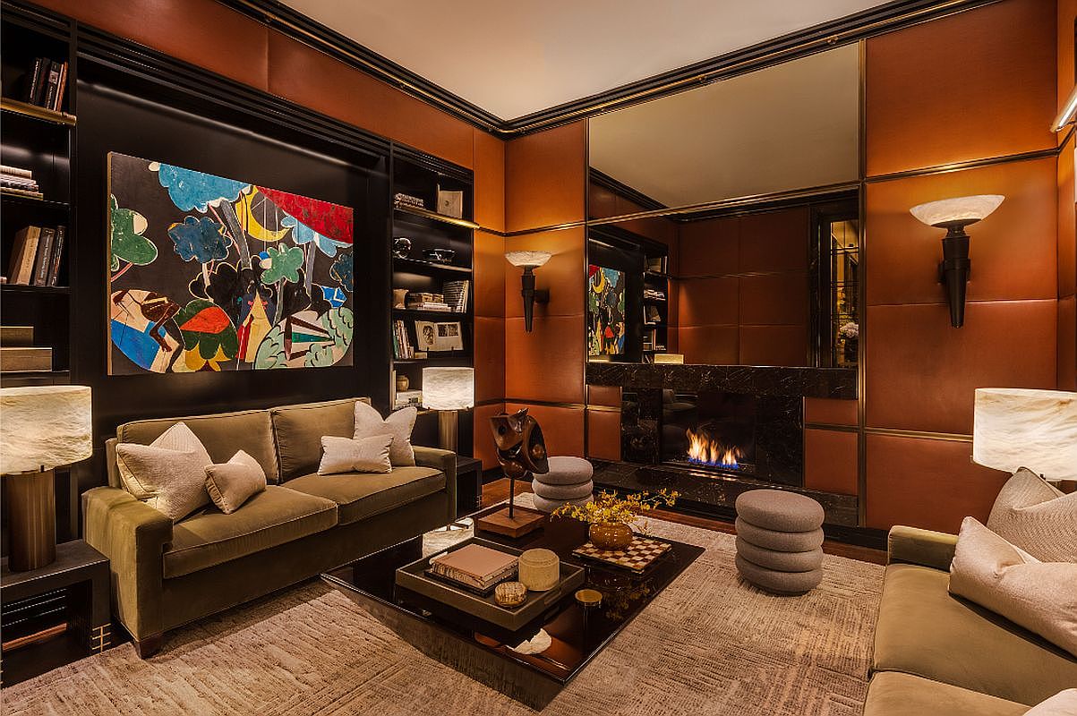 This is a luxurious living room featuring a warm color palette with brown leather paneled walls and black built-in shelving. The room is furnished with two olive green sofas, a dark glass coffee table, and a fireplace with a mirror above it. An abstract painting adds a pop of color to the space, creating a sophisticated and inviting atmosphere.