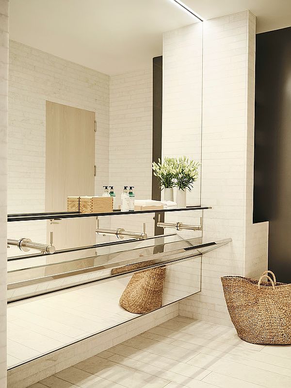 This is a modern bathroom featuring white brick walls and light-colored tile flooring. A long, mirrored vanity with a glass shelf underneath provides ample storage space, with woven baskets adding a touch of natural texture. The room is well-lit and appears clean and minimalist, creating a serene and inviting atmosphere.