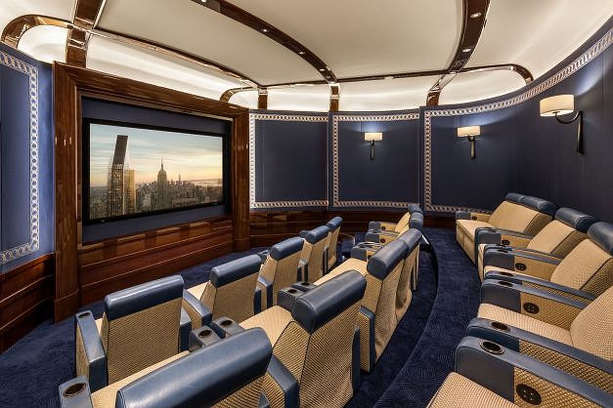 This is a luxurious home theater featuring tiered seating with plush, leather-accented chairs and integrated cup holders. A large screen displays a cityscape, framed by rich wood paneling and elegant wall sconces. The room is designed for an immersive cinematic experience with a curved layout and sophisticated lighting.