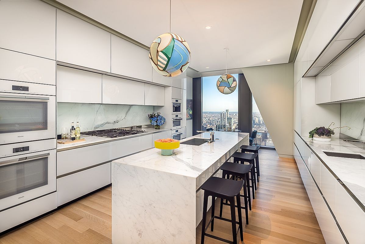 This is a modern, high-end kitchen featuring sleek white cabinetry, marble countertops, and stainless steel appliances. A large island with bar seating anchors the space, while unique pendant lighting adds a touch of artistry. The kitchen boasts a stunning city view through a large window, enhancing the luxurious feel.