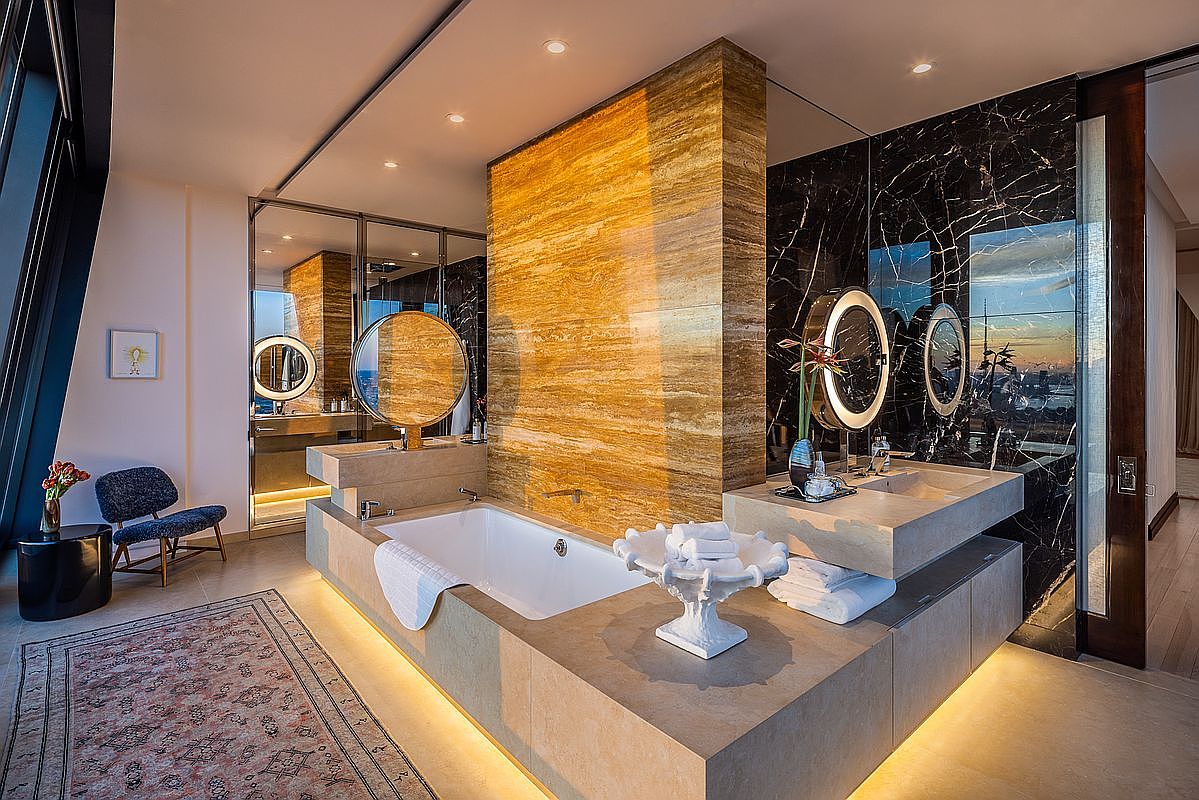 This is a luxurious primary bathroom featuring a modern design with high-end finishes. The bathroom includes a freestanding bathtub surrounded by a beige stone platform with underlighting, a separate vanity area with dual sinks and round mirrors, and black marble wall accents. A large window offers a view of the city skyline, enhancing the spa-like atmosphere.