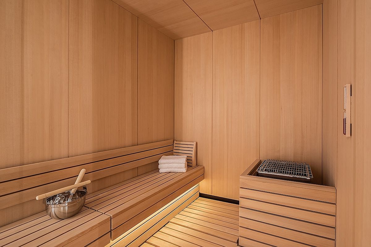 This is an interior shot of a sauna, featuring wooden walls, benches, and flooring. A metal bucket with wooden dippers sits on the lower bench, and a stack of white towels is neatly arranged on the upper bench. The sauna heater is visible in the corner, creating a warm and inviting atmosphere.