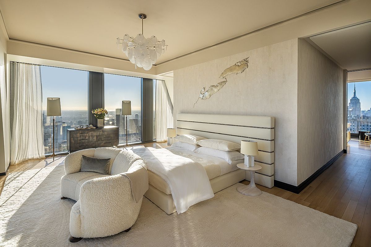 This is a luxurious primary bedroom with a large bed, a comfortable armchair, and floor-to-ceiling windows offering stunning city views. The room features a neutral color palette, a modern chandelier, and an elegant accent wall, creating a serene and sophisticated atmosphere. The perspective is from the doorway, showcasing the entire room.