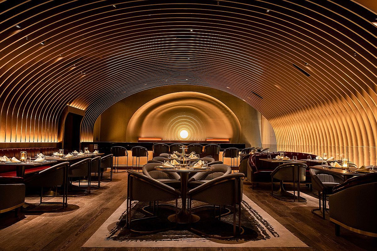 This is a stunning dining room with a unique architectural design featuring a curved, ribbed ceiling and a focal point at the back. The room is furnished with elegant tables and chairs, creating a sophisticated and intimate dining atmosphere. The warm lighting enhances the ambiance, making it an ideal space for upscale dining experiences.