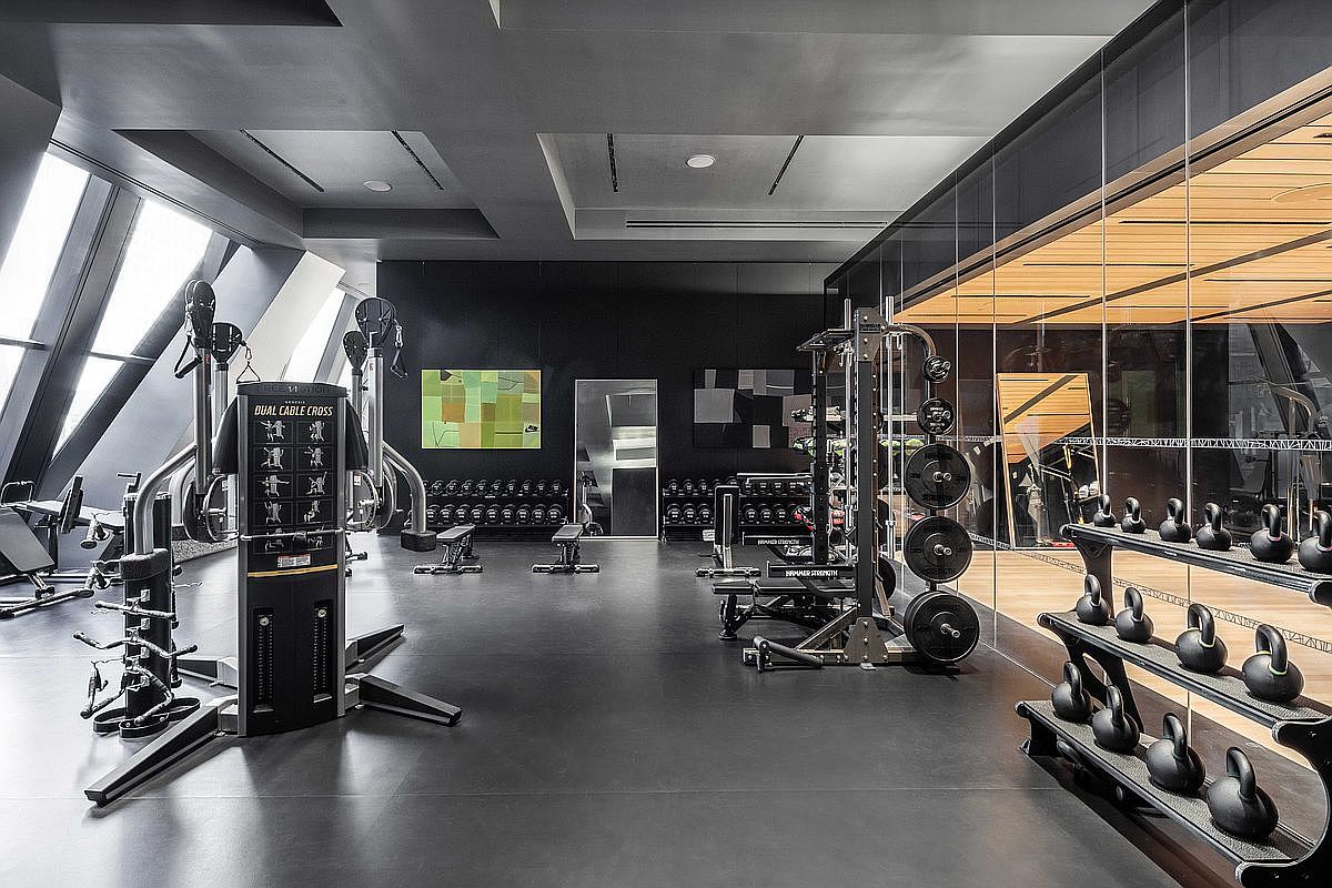 This is a modern home gym featuring a variety of exercise equipment, including a dual cable cross machine, weight racks, dumbbells, and kettlebells. The room has a sleek, contemporary design with dark walls, black flooring, and large windows that provide natural light. The space is well-organized and appears to be in a luxury residential setting.