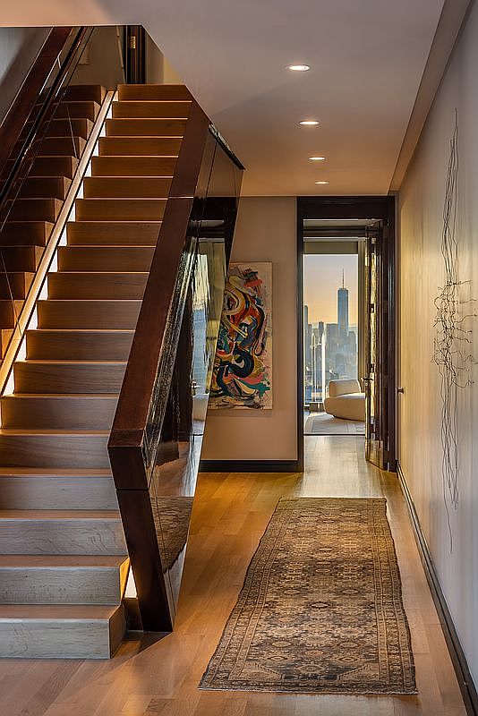 This interior shot showcases a modern hallway with wooden stairs and a dark wood railing. An abstract painting hangs on the wall, and a long rug stretches down the hallway. A doorway at the end of the hall reveals a stunning city view, adding a touch of luxury and sophistication to the space.
