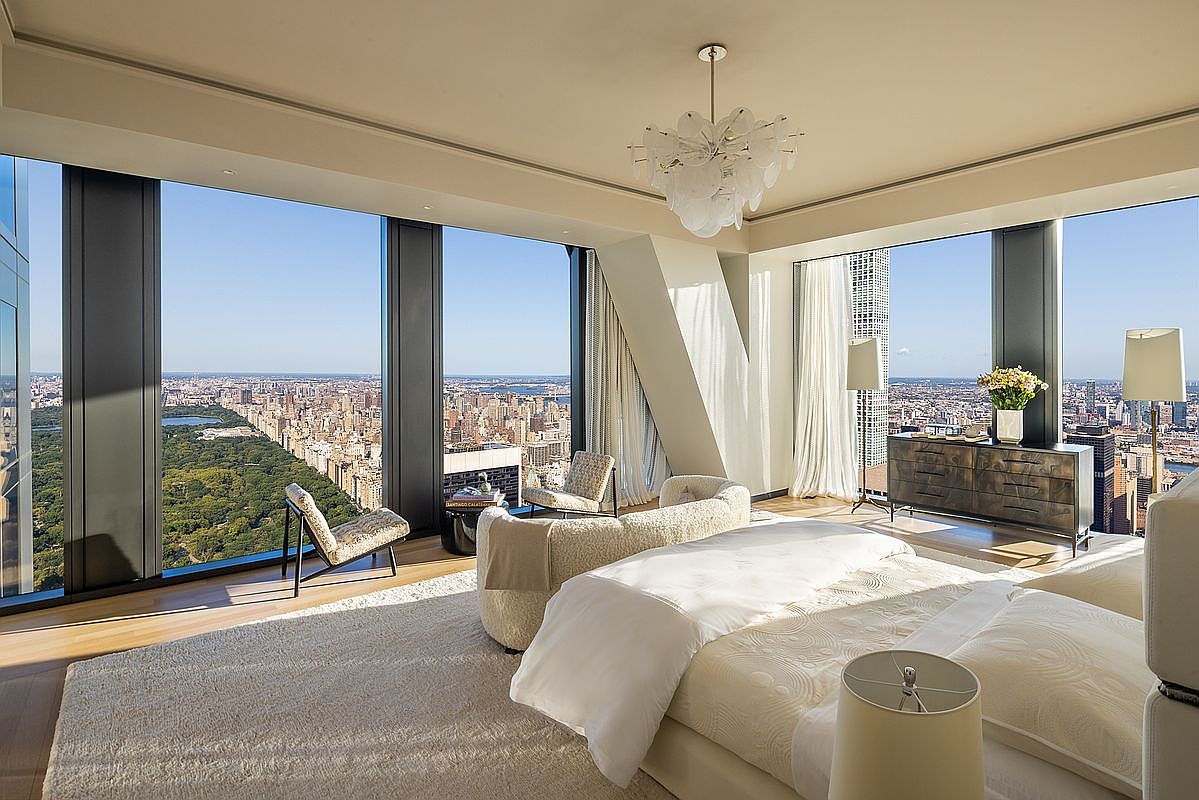 This is a luxurious primary bedroom with floor-to-ceiling windows offering panoramic city views. The room features a large bed with white linens, a plush rug, and stylish seating areas. A modern chandelier hangs from the ceiling, and a sleek dresser adds to the room's sophisticated ambiance.