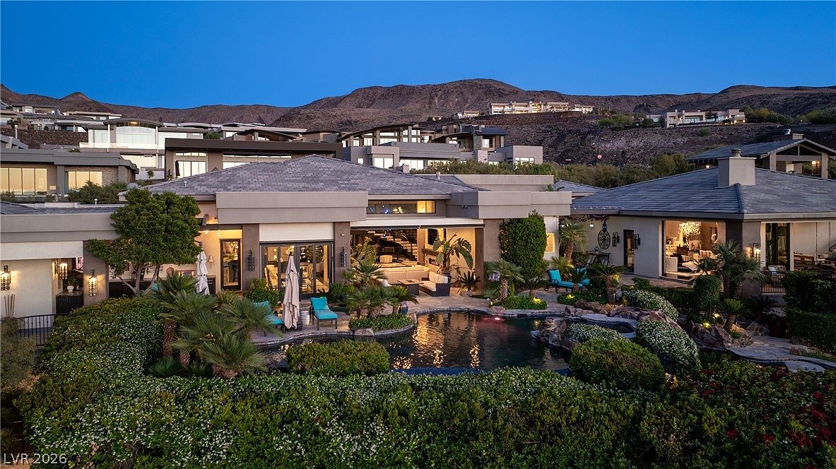 This expansive rear view captures a luxurious modern residence at dusk, featuring a multi-level design with a central swimming pool and integrated spa. The home showcases large glass sliding doors that open to a covered patio area, creating a seamless transition between indoor and outdoor living spaces. Lush landscaping, including manicured hedges and palm trees, surrounds the pool, while the backdrop features a dramatic mountain range under a twilight sky.