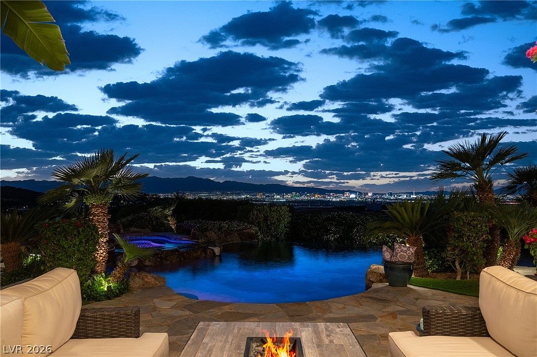 This evening view from a stone-paved patio captures a luxurious outdoor living space featuring a fire pit in the foreground and a tranquil swimming pool. Beyond the pool, lush landscaping frames a panoramic, sparkling city skyline set against a dramatic, cloud-filled twilight sky. The perspective is from a seated position, emphasizing the relaxing atmosphere and the seamless transition between the private terrace and the expansive horizon.