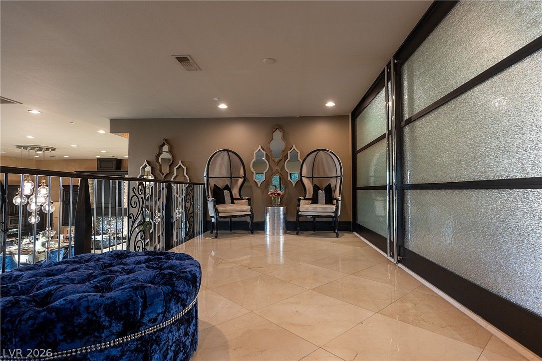 This elegant landing area features a polished light-toned tile floor and a prominent blue velvet tufted ottoman in the foreground. The space is accented by two high-backed, wing-style chairs flanking a small metallic side table, set against a wall decorated with a cluster of ornate, decorative mirrors. To the right, a large, multi-panel glass partition with a dark frame provides a modern architectural element, while a wrought-iron railing overlooks the lower level of the home.