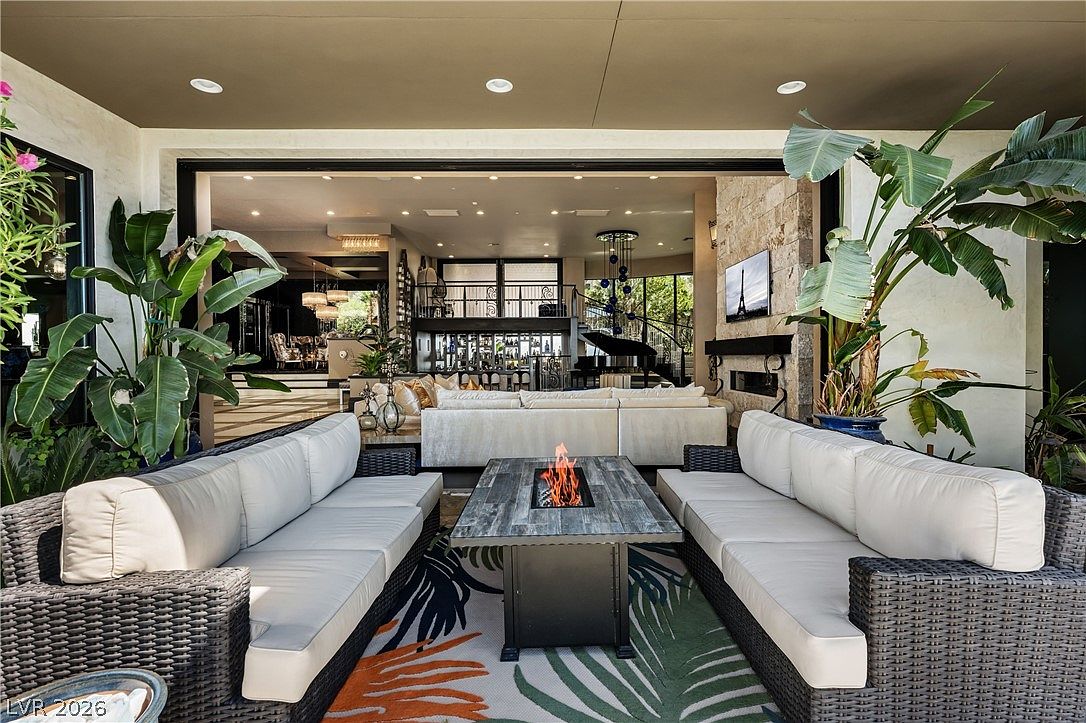 This luxurious covered patio features a sophisticated outdoor living area with two large, dark-wicker sofas facing a central fire pit table. The space seamlessly transitions into an expansive indoor living room through a wide, open-wall design, showcasing a grand interior with a piano and bar area. Lush potted plants frame the scene, creating a resort-like atmosphere perfect for high-end entertaining.