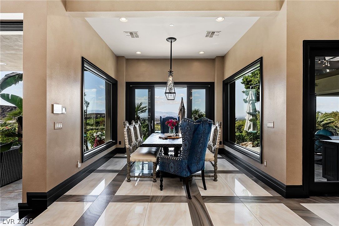 This elegant dining room features a formal table set with ornate, blue-upholstered chairs, centered beneath a decorative pendant light. Large windows on either side of the room provide expansive views of the outdoors, while the floor is finished with a sophisticated pattern of light-toned tiles and dark borders. The space is framed by dark trim and neutral-toned walls, creating a refined and balanced atmosphere.
