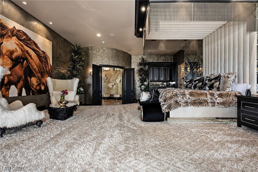 This opulent primary bedroom features a plush, high-pile carpet and a large bed adorned with a textured animal-print throw. A striking, oversized horse portrait hangs on the wall above a pair of white accent chairs, while a dark, ornate cabinet and a metallic chain-link ceiling feature add to the dramatic, high-end aesthetic. The room is framed by double doors leading to an adjacent space, creating a sense of grandeur and sophisticated luxury.