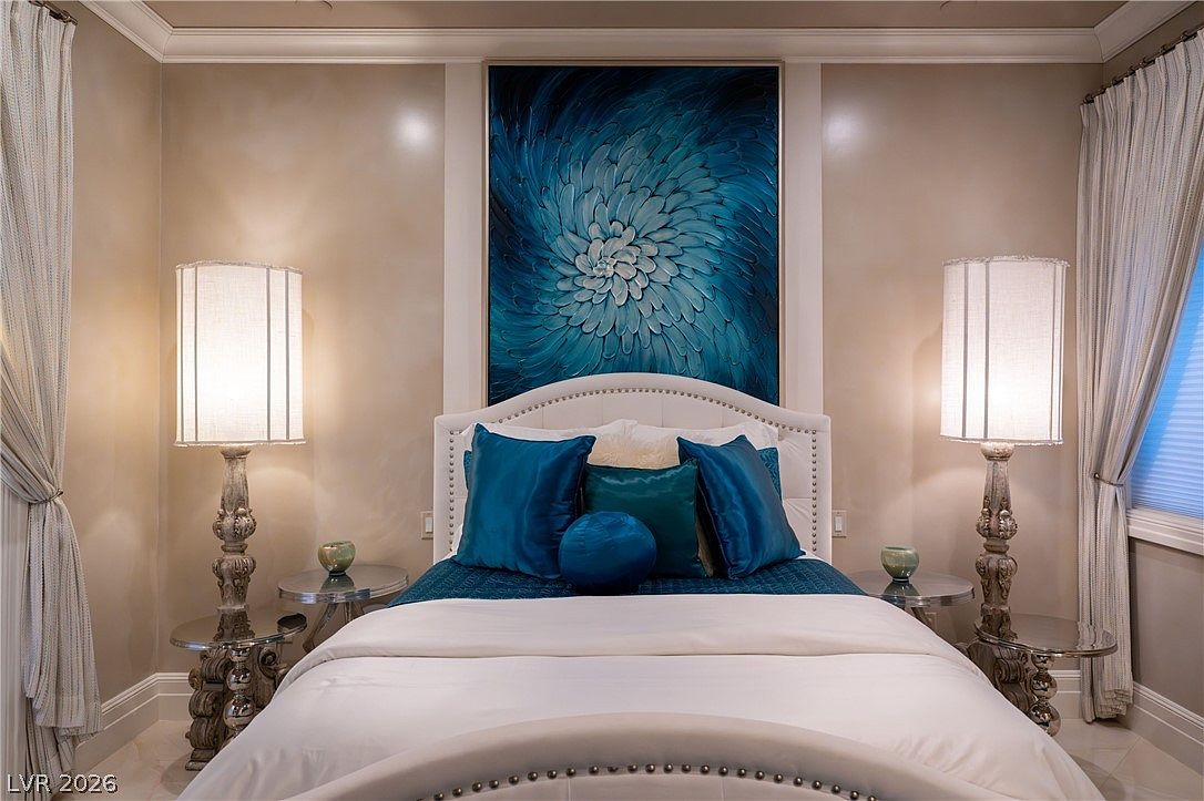 This bedroom features a symmetrical layout centered around a white upholstered headboard and a large, vibrant blue floral abstract painting. Flanking the bed are two matching metallic side tables topped with tall, textured lamps that provide soft, ambient lighting. The room is framed by elegant floor-to-ceiling curtains on either side, creating a sophisticated and tranquil atmosphere.
