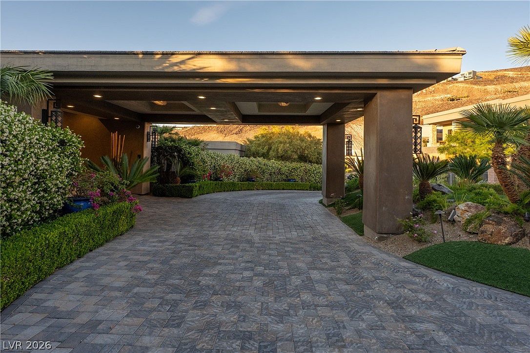 This image captures a grand, covered porte-cochère entrance to a luxury residence, featuring a paved driveway with intricate stone patterns. The structure is supported by substantial pillars and includes recessed lighting, creating a sophisticated and welcoming arrival experience. Lush landscaping, including manicured hedges and palm trees, frames the driveway, while the backdrop reveals a desert hillside setting.