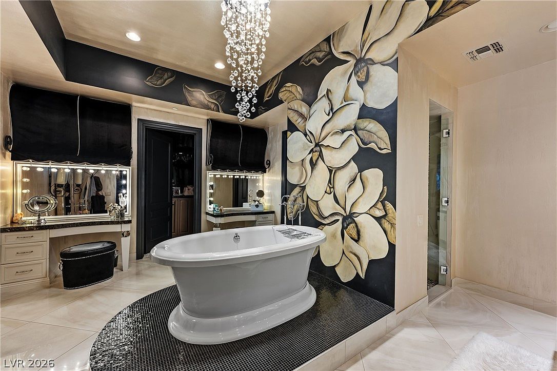 This luxurious primary bathroom features a central freestanding soaking tub set upon a dark, circular mosaic-tiled platform. A dramatic, large-scale floral mural adorns the wall behind the tub, complemented by a sparkling crystal chandelier and dual vanity stations with illuminated mirrors. The space is finished with light-toned tile flooring and a glass-enclosed shower, creating an opulent and sophisticated atmosphere.